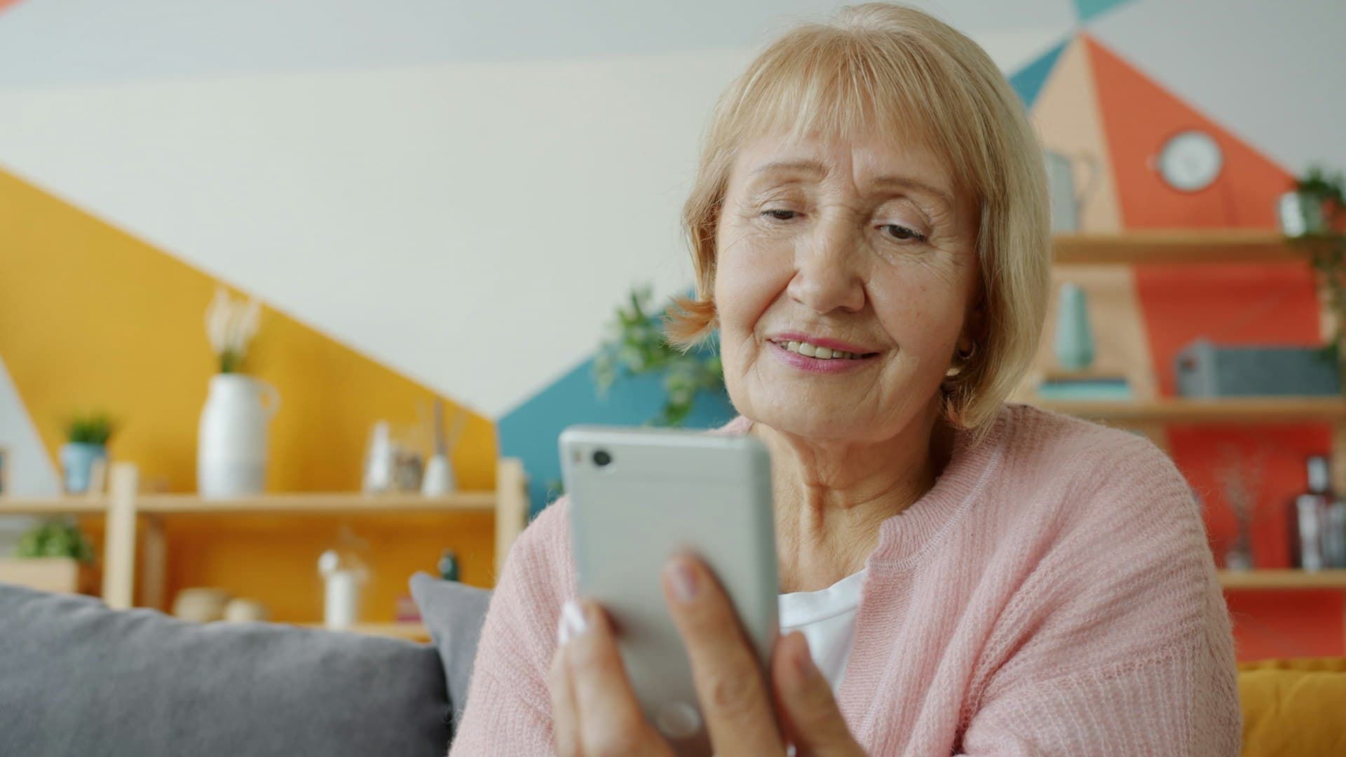 An older woman in a pink cardigan smiles at her phone at home.